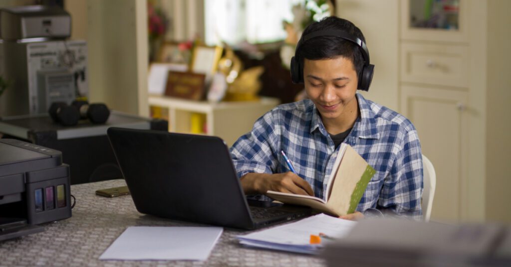 A male teenage student wearing headphones participates in an online class from home, using his laptop and taking notes with pen and paper